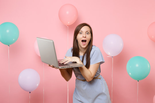 Portrait of shocked young woman with opened mouth in blue dress holding using laptop pc computer on pink background with colorful air balloons. Birthday holiday party people sincere emotions concept.