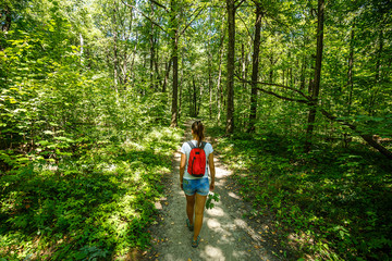 Young girl backpacker in jeans shorts with red backpack and sandals on his feet, walks on the road in the green forest in the sunshine outdoors in the summer time