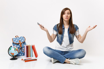 Young shocked puzzled woman student holding mobile phone, spreading hand, sitting near globe, backpack, school books isolated on white background. Education in high school university college concept.