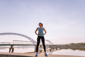 Confident strong cute sporty blond girl standing on jetty near river after early morning training.