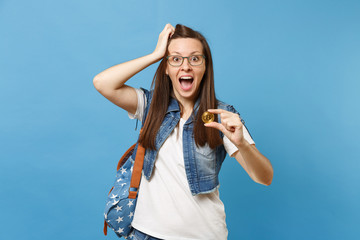 Young amazed shocked woman student with opened mouth clinging to head hold bitcoin metal coin of golden color isolated on blue background. Future currency. Education in high school university college.