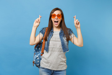 Young excited woman student with opened mouth wearing orange heart glasses waiting for special moment, keeping fingers crossed isolated on blue background. Education in high school. Advertising area.