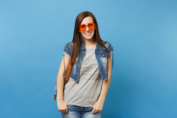 Portrait of young attractive woman student in denim clothes with backpack wearing orange heart glasses standing isolated on blue background. Education in high school. Copy space for advertisement.