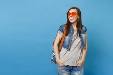 Young joyful cute woman student with backpack wearing orange heart glasses looking aside holding hands in pockets isolated on blue background. Education in high school. Copy space for advertisement.