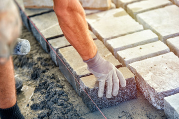 A workman's gloved hands use a hammer to place stone pavers. Worker creating pavement using...