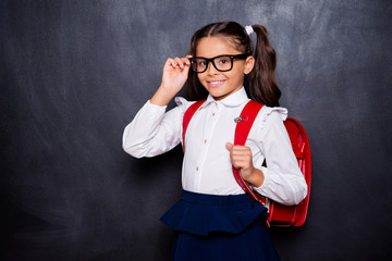 Portrait of nice smart cute stylish adorable small little girl with curly ponytails in white blouse shirt and blue skirt with bag, touching glasses. Isolated over black background