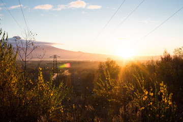 Sunrise is during great morning in mountains. Big metal high-voltage line is in right side. Beautiful grass and trees are under warm light. Sky is highlighted by the sun. Golden autumn in mountains.