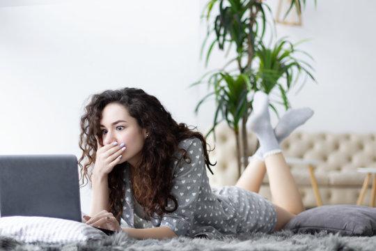 Girl female woman wearing pajamas sleepwear with white hearts lying on the carpet in light interior room and happy smiling working with laptop. Freelancer, working at home, success, surprized concept.