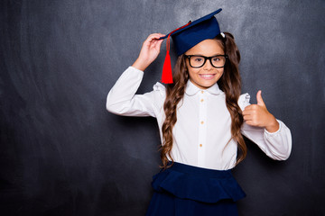 Choose great international education abroad! Positive glad nice adorable cute small little girl with curly ponytail in white shirt, skirt, master's hat, giving thumb-up. Isolated over black background