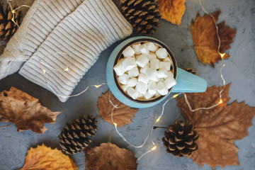 Blue mug with coffee, hot chocolate or cocoa with marshmallow on gray concrete background and lying dry leaves, cones, sweater, garland. Concept of warm cozy autumn or winter.