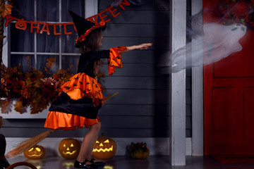 Young funny girl kid child in halloween orange costume of witch and black hat playing outdoor with witch broom and spooky jack lantern pumpkins with scary faces and chasing ghost. Halloween concept.