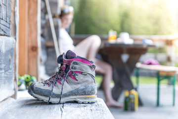 Paar Bergschuhe auf Alm, hölzerne Veranda, Tisch mit Wanderer im Hintergrund