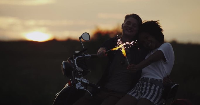 Charismatic young couple sitting on retro bike at sunset holding the sparklers and smiling holding each other.
