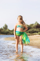 little joyful blonde with a tail girl in a swimsuit and a pareo posing and jumping on the river bank in sunny summer weather