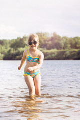 a small blonde girl with a tail in a swimsuit and glasses fooling around jumping splashes in the sunny warm summer weather in the river