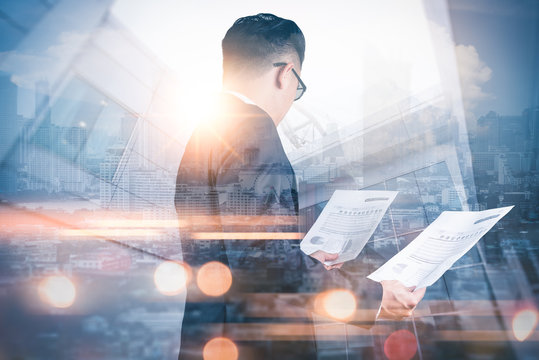 The double exposure image of the businessman standing and looking to documents during sunrise overlay with cityscape image. The concept of modern life, business, city life and internet of things.