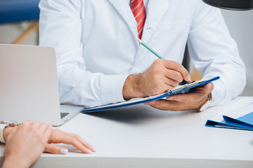 partial view of physiotherapist in white coat making notes in notepad with female patient near by...
