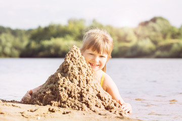 a small blonde joyful girl in a swimsuit builds a sand castle on the river bank in sunny warm weather in the summer. portrait
