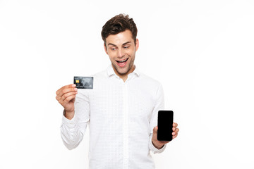 Happy guy with dark hair holding mobile phone and credit card, isolated over white background