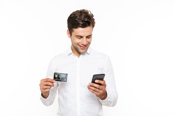 Excited young man with dark hair holding smartphone and credit card, isolated over white background