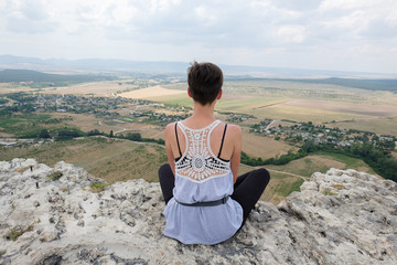 young woman sitting on peak of a cliff and looking forward to the plain. Female hiker  on top of mountain and enjoying view. Travel, freedom, vacation, trekking, hiking, zen concept