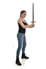 full length portrait of brunette girl wearing black single and jeans. standing pose holding a sword. isolated on white studio background.