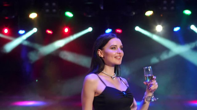 Portrait of a sexy girl in a long evening dress with champagne in the dark under the lights of bright spotlights. Young girl with a glass of champagne dancing at a party in a nightclub, slow motion.