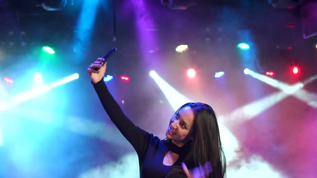 Portrait of happy girl in evening dress with a telephone on a party. Funny girl dancing in a nightclub and makes a selfie. Slow motion. Dark. Multi-colored spotlights.