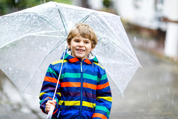 Beautiful little kid boy on way to school walking during sleet, rain and snow with an umbrella on cold day. Happy and joyful child in colorful fashion casual clothes