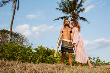 Smiling girlfriend hugging boyfriend with surfboard near palm trees in Bali, Indonesia