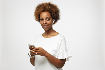Studio photo of young african lady isolated on gray background standing with positive smile, holding cellphone in hands, wearing smartwatch and wireless earphones, feeling confident