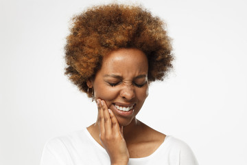 Young african american woman in blank white t shirt, isolated on gray background, touching her face with expression of horrible suffer from health problem and aching tooth