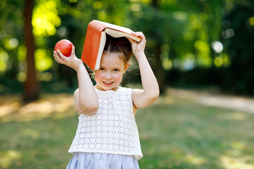 Funny adorable little kid girl with book, apple and backpack on first day to school or nursery. Child outdoors on warm sunny day, Back to school concept. Healthy child of elementary class.