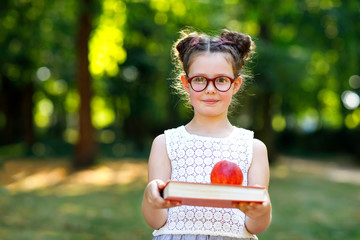 Funny adorable little kid girl with glasses, book, apple and backpack on first day to school or nursery. Child outdoors on warm sunny day, Back to school concept. Healthy child of elementary class.