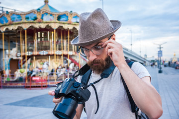 bearded young man in hat and eyeglasses using digital camera in amusement park