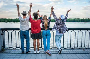 Obraz premium back view of four multiethnic friends raising hands at riverbank