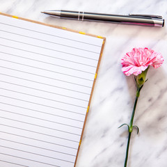 Naklejka premium A square photo of a marble desk seen from above with an open lined notebook, a pen and a pink carnation