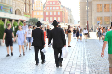 Dwóch mężczyzn w czarnych kapeluszach i garniturach idą z reklamówkami przez miasto.