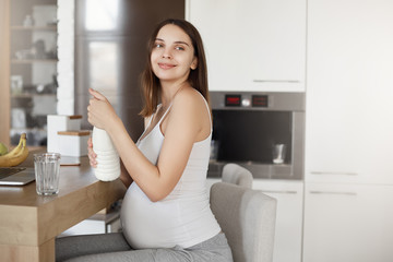 Fototapeta na wymiar Keeping water balance is important. Indoor shot of charming carefree future mother, being pregnant, sitting in kitchen near laptop, pouring milk in glass and gazing aside with warm smile
