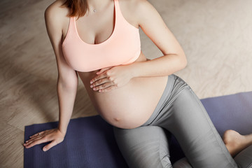 Fototapeta na wymiar Cropped shot of tender caring pregnant female in leggings and pink sports bra, touching belly while thinking about future baby be born, lying on roll pad at home, taking rest after breathing exercises