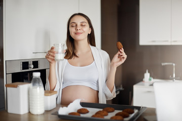 Pregnant woman feeling delight and joy, enjoying great taste of fresh baked cookies and drinking warm milk, closing eyes and smiling, holding glass and cookie, sitting near kitchen table