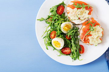 Tasty breakfast. Open sandwiches with salmon, cream cheese and rye bread in a white plate and salad with tomato, egg and arugula. Top view. Flat lay