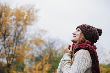 Model wearing warm winter hat, sweater and red scarf and looking up on the sky. The girl folded her arms together on the chest. Woman holding her hands on the scarf. Outdoor. Profile portrait. © darakaliton