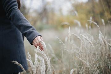 Woman in grey overcoat walking on the field and touching spikelets. 