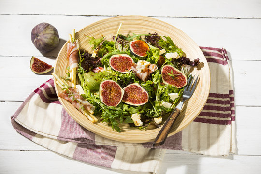gemischter Blattsalat mit Feigen, Tomaten, Schafsk&auml;se und Crossinis mit Schinken,serviert auf Holzteller, weisser Holztisch 