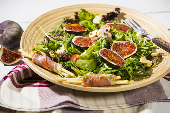 gemischter Blattsalat mit Feigen, Tomaten, Schafsk&auml;se und Crossinis mit Schinken,serviert auf Holzteller, weisser Holztisch 