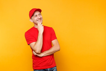 Delivery man in red uniform isolated on yellow orange background. Professional smiling male employee in cap, t-shirt working as courier or dealer put hand prop up on chin. Service concept. Copy space.