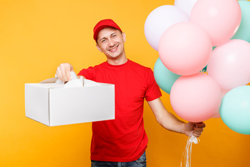 Man giving food order cake box isolated on yellow background. Male employee courier in red cap t-shirt hold colorful air balloons, dessert in empty cardboard box. Delivery service concept. Copy space.