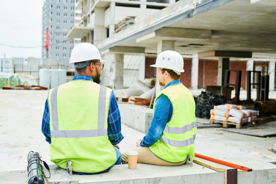 Back view portrait of two workers wearing hardhats  chatting while enjoying coffee break on construction site