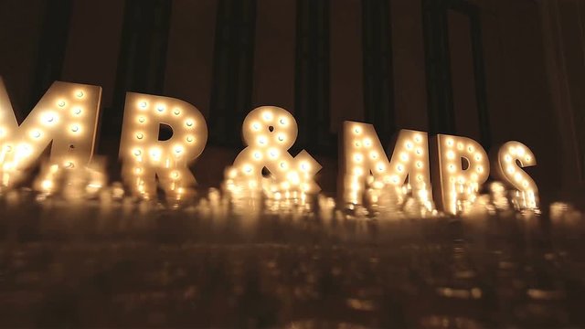 Retro lIghtbox theater style of text Mr & Mrs decoration on the backdrop in the wedding recepton dinner night party with a group of wine glasses blurry in foreground, Low angle, Dolly slider scene.